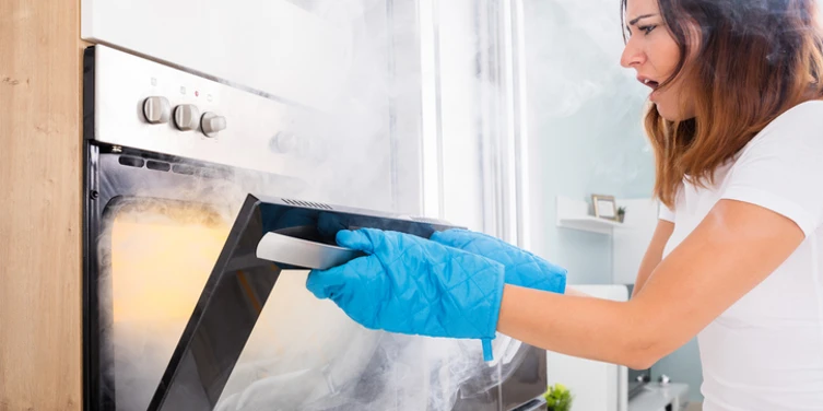 A woman opens an oven with concern on her face and smoke rising out of the appliance.