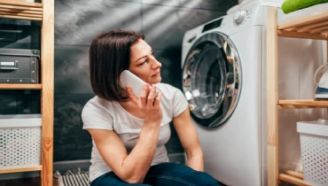 Woman sitting beside her washing machine on the phone, looking disappointed.