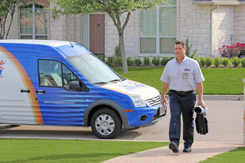 Service technician carrying a tool bag and walking up a sidewalk with work van parked on the street behind him.